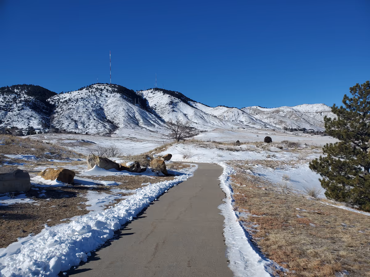 A concrete path leads down a hill. There is snow on either side, and mountains are in the background.