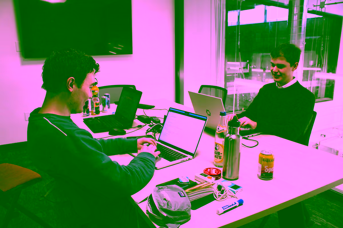 Lukas and Grant write code on their laptops. Beverage cans, electronics, and miscellaneous paraphernalia are strewn across their table.